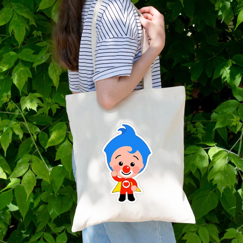 May include: A cream-colored canvas tote bag featuring a graphic print of a cartoon character with bright blue hair, a red nose, a red shirt with a white heart emblem, black pants, and a yellow cape, positioned centrally on the front of the bag.