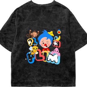 May include: A black acid-wash t-shirt featuring a colorful graphic print of the character Plim Plim with a monkey, a bee, a spider, a chick, and a banana. The text Plim Plim is displayed in large, stylized, multi-colored bubble letters.