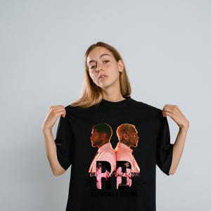 May include: A black t-shirt featuring a graphic print of two individuals in pink suits facing away from each other. The text reads THE R&B TOUR and RAYMOND & BROWN, with smaller images of the individuals positioned within the large letters R&B.