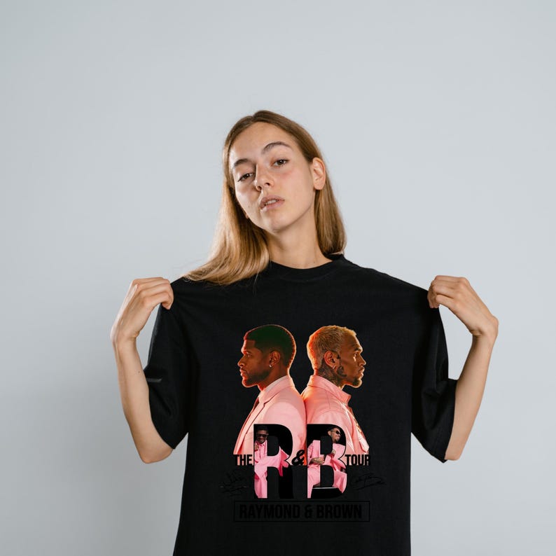 May include: A black t-shirt featuring a graphic print of two individuals in pink suits facing away from each other. The text reads THE R&B TOUR and RAYMOND & BROWN, with smaller images of the individuals positioned within the large letters R&B.