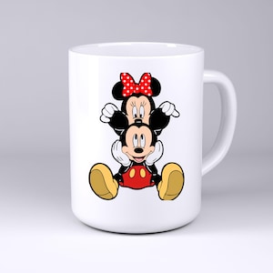 May include: A white ceramic mug featuring a graphic illustration of Minnie Mouse sitting on top of Mickey Mouse. Minnie wears a red bow with white polka dots, while Mickey is depicted with his signature red shorts and large yellow shoes.