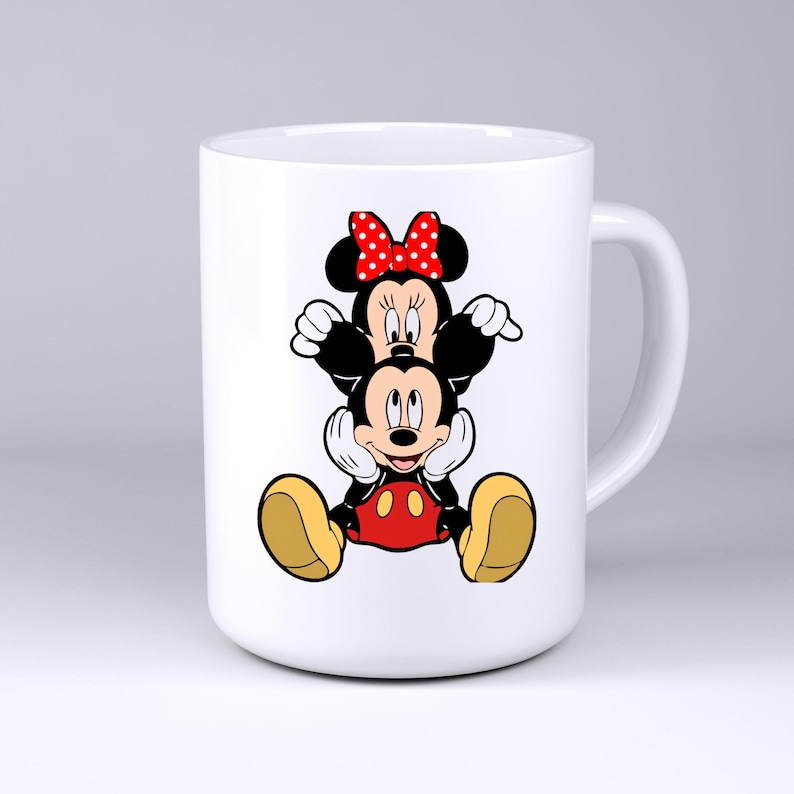 May include: A white ceramic mug featuring a graphic illustration of Minnie Mouse sitting on top of Mickey Mouse. Minnie wears a red bow with white polka dots, while Mickey is depicted with his signature red shorts and large yellow shoes.