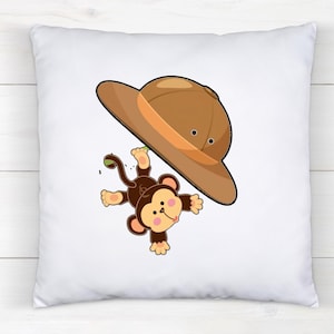 May include: A square white throw pillow featuring a cartoon illustration of a brown monkey hanging from a tan safari hat. The monkey has a light brown face, pink cheeks, and a long tail. The design is centered on the front of the pillow cover.