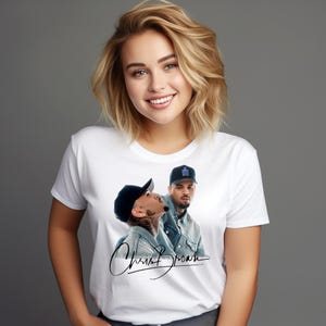 May include: A white short-sleeved t-shirt featuring a graphic print of Chris Brown wearing a denim jacket and a baseball cap. Below the image of the artist is a black cursive signature that reads Chris Brown. The shirt has a crew neckline.