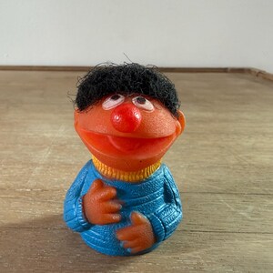 May include: A molded rubber figurine of a character with orange skin, a round red nose, and black hair. The figure wears a blue textured sweater with a yellow collar. The character has wide eyes looking upward and an open mouth with a visible tongue.