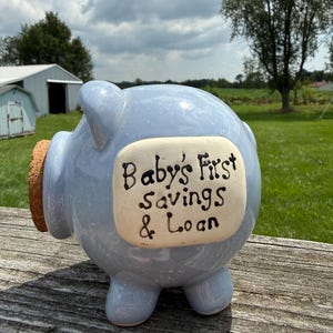 May include: A light blue ceramic piggy bank with a glossy finish and a cork stopper in the snout. A rectangular cream-colored plaque on the side features the hand-painted black text "Baby's First Savings & Loan" in a whimsical script font.