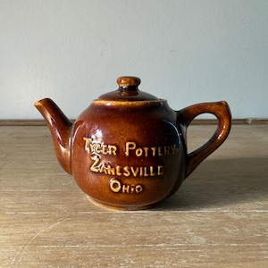 May include: A brown glazed ceramic teapot with a lid, a curved handle, and a spout. The front of the teapot features embossed text that reads Tyger Pottery Zanesville Ohio in a light brown colour, contrasting with the dark brown glossy finish.