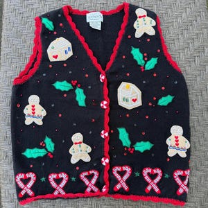 May include: A black knit vest featuring a red scalloped trim and peppermint-style buttons. The vest is decorated with appliqués of gingerbread people, gingerbread houses, green holly leaves, and red candy canes along the bottom hem.