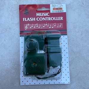 May include: A dark green music flash controller device encased in clear plastic packaging. The backing card is red with white text reading MUSIC FLASH CONTROLLER and a musical staff graphic. The bottom section has a white background with red dots.