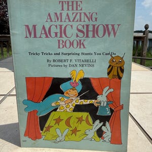 May include: A light blue hardback book titled The Amazing Magic Show Book by Robert F. Vitarelli with pictures by Dan Nevins. The cover features a cartoon illustration of a magician with a wand, three rabbits, and an owl on a stage.