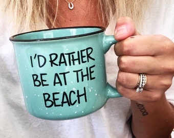 May include: A turquoise blue ceramic mug with white speckles and a black rim. The mug has the text "I'd rather be at the beach" printed in black.