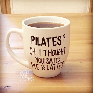 May include: White ceramic mug filled with a dark liquid, possibly coffee or tea. The mug has a large handle and features the text "PILATES? OH I THOUGHT YOU SAID PIE & LATTES" in black lettering. The words "SIMPLY MADE GREETINGS" are also printed on the mug.