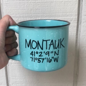 May include: A light blue ceramic mug with a black rim and white speckles. It says "Montauk" in black lettering and the coordinates "41°2'9"N 71°57'16"W" in black lettering.