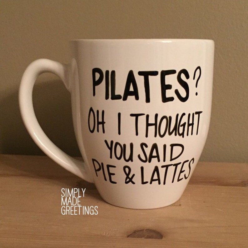 May include: White ceramic mug with a curved handle. The mug features black text that reads "PILATES? OH I THOUGHT YOU SAID PIE & LATTES." The words "SIMPLY MADE GREETINGS" are printed at the bottom.