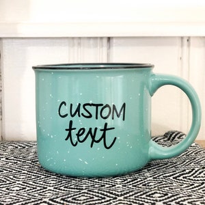 May include: A light blue ceramic mug with a black rim and a speckled glaze. The mug has black text that says "CUSTOM text".