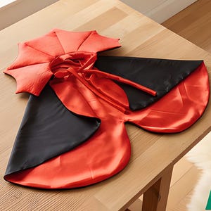 May include: A reversible cape featuring a vibrant red satin side and a solid black satin side. The garment includes a structured, pleated collar and long fabric ties at the neck for fastening. The design is smooth, lightweight, and glossy.