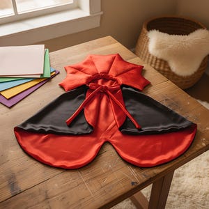 May include: A reversible cape featuring a vibrant red satin side and a solid black satin side. The garment includes a structured, pleated red collar and long red fabric ties at the neck for closure. The cape is laid flat, displaying its smooth texture.