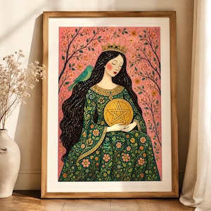 May include: A framed art print featuring a long-haired figure wearing a crown and a green floral dress, holding a golden pentacle. The background is a pink landscape with flowering branches and a small blue bird perched on the figure's shoulder.