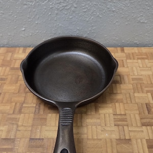 May include: A dark brown cast iron skillet with a smooth interior surface, a rounded handle featuring a teardrop-shaped hole, and a ribbed thumb rest. A white tag attached to the handle reads Benjamin & Medwin 6.5 in Skillet Sanded.