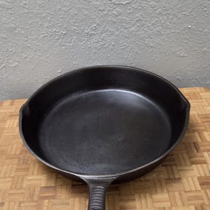 May include: A black cast iron skillet with a round cooking surface, a long handle featuring a ribbed thumb rest, and a hole at the end. A white tag attached to the handle displays the text: Unmarked Benjamin & Medwin 10 in Sanded - $20.