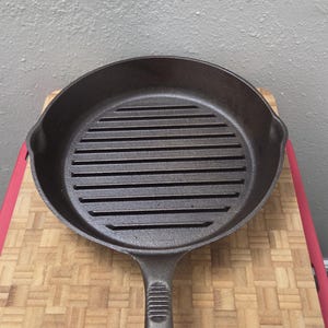May include: A round, dark brown cast iron grill pan featuring a textured interior surface with horizontal raised ridges for searing. The pan includes a long, sturdy handle with a ribbed thumb grip and a small hole at the end for hanging storage.