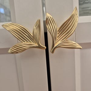 May include: Two gold-coloured metal cabinet pulls shaped like stylised leaves with prominent, raised linear veins. Each handle features a central stem branching into three distinct, pointed leaf segments with a smooth, matte metallic finish.