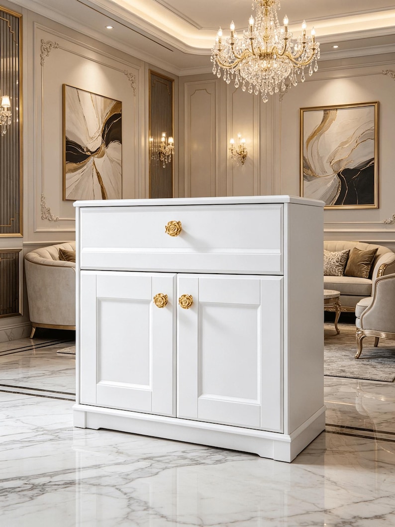 May include: A white cabinet featuring a single top drawer and two lower doors, each equipped with a gold-colored rose-shaped knob. The cabinet has a rectangular frame with recessed panel detailing on the drawer front and the two cabinet doors.
