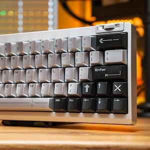 May include: A mechanical keyboard with a silver metallic frame and a two-tone colour scheme. The alphanumeric keys are light grey, while the modifier keys are black. A silver rotary knob is positioned at the top right corner of the frame.