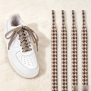 May include: A white low-top sneaker featuring a perforated toe box and a padded collar, laced with flat shoelaces in a brown and beige houndstooth pattern. Beside the sneaker are four additional matching houndstooth patterned shoelaces.