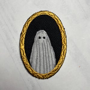 Può includere: Una toppa ricamata di forma ovale raffigurante una figura di fantasma grigio chiaro con due piccoli occhi neri centrati su uno sfondo nero solido, il tutto racchiuso in un bordo decorativo color oro metallizzato con un motivo testurizzato.