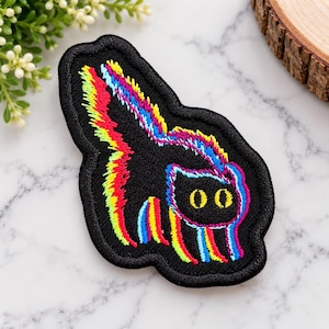 Puede incluir: Un parche bordado que presenta la silueta de un gato negro con acentos de hilo multicolor vibrante en amarillo, rojo y azul a lo largo de sus bordes y cola. El gato tiene dos ojos de color amarillo brillante y una postura arqueada.