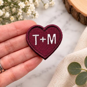 May include: A heart-shaped embroidered patch in a deep burgundy color featuring the white stitched letters T + M in the center. The patch has a thick, matching burgundy border and a textured fabric surface, showcasing clean embroidery work.