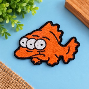 May include: An embroidered patch shaped like a three-eyed orange fish with a black border. The fish has a jagged dorsal fin, a wide tail, and three white circular eyes with black pupils arranged horizontally across its orange body.