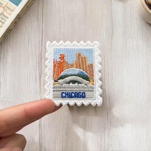 Puede incluir: Un parche bordado rectangular con forma de sello postal y borde blanco festoneado. El diseño muestra el horizonte de Chicago en tonos naranja y beige, una estructura de cúpula azul y blanca, y la palabra CHICAGO en hilo azul.