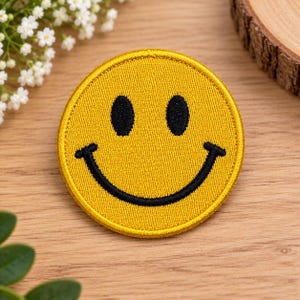 Peut inclure: Un écusson brodé circulaire présentant un fond jaune vif avec un motif de visage souriant cousu en noir. L'écusson possède une bordure jaune épaisse et une surface en tissu texturé, affichant deux yeux ovales noirs et une large bouche incurvée.