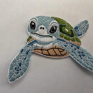 Puede incluir: Un parche bordado que representa una tortuga marina con cabeza azul claro, grandes ojos negros y un caparazón con patrones en tonos verdes y bronceados. La tortuga tiene aletas azul claro con detalles en azul oscuro y un borde blanco.