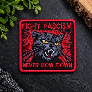 Puede incluir: Un parche bordado cuadrado con borde negro y ribete rojo. Presenta un gato negro con ojos amarillos y la boca abierta, rodeado de líneas rojas radiantes. El texto FIGHT FASCISM está en la parte superior y NEVER BOW DOWN en la inferior.