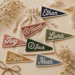 May include: A collection of triangular felt pennants featuring embroidered names including Ethan, Hazel, Ivy, Luna, Jack, Piper, and Noah. Each pennant displays a unique color, some with scalloped edges or circular monograms, arranged around a drawstring bag.