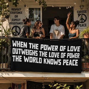 May include: A black horizontal banner featuring the white text: WHEN THE POWER OF LOVE OUTWEIGHS THE LOVE OF POWER THE WORLD KNOWS PEACE. The banner is displayed on a balcony railing, with four people standing behind it in a sunlit outdoor setting.