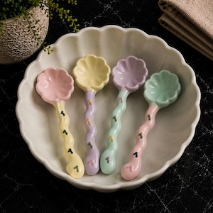 May include: Four ceramic spoons with flower-shaped bowls and wavy handles in pastel pink, yellow, lavender, and mint green. Each handle features small, colourful, scattered dash-like patterns. The spoons are arranged inside a white scalloped bowl.