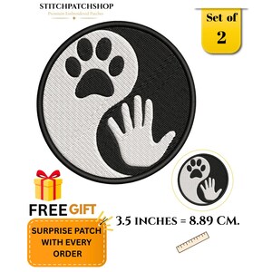 May include: A circular embroidered patch featuring a black and white yin-yang design with a paw print and a human hand silhouette. The patch measures 3.5 inches in diameter. The set includes two patches with a black border and detailed stitching.