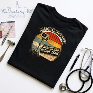 Puede incluir: Una camiseta negra con un gráfico de un escáner de vejiga médico dentro de un círculo de atardecer desgastado. El texto dice BLADDER SCANNER SEARCH AND RESCUE TEAM. El diseño utiliza colores naranja, amarillo y azul con textura vintage.