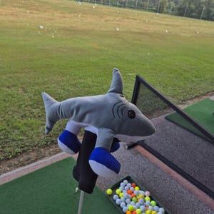 May include: A plush shark golf club headcover featuring a grey body, white underbelly, and black eyes. The shark has two blue and white fins extending from the sides and is attached to a black fabric sleeve that fits over a golf club shaft.