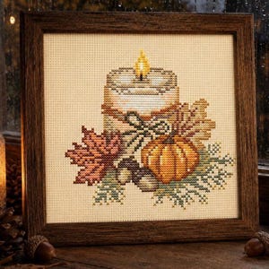 May include: A framed cross-stitch embroidery featuring a lit candle with a yellow flame, a small orange pumpkin, and autumn leaves in shades of red, brown, and green. The design is centered on a light beige fabric within a dark brown wooden frame.