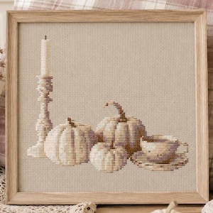 Könnte beinhalten: Ein gerahmtes Kreuzstich-Stickbild mit einer hohen Kerze im Ständer, drei Kürbissen unterschiedlicher Größe und einer Teetasse auf einer Untertasse. Das Design verwendet neutrale Beige-, Creme- und hellbraune Töne auf einem hellen Stoffhintergrund.