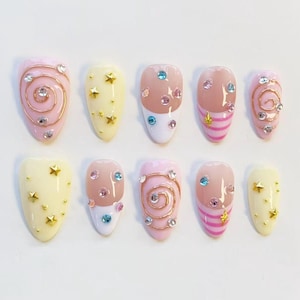 May include: A set of ten press-on nails featuring a variety of designs including pale pink and yellow bases, gold wire swirls, small gold star studs, pink horizontal stripes, and embedded multicoloured rhinestones in various circular shapes.
