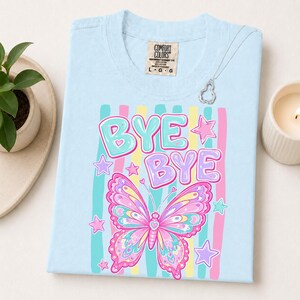 May include: A light blue t-shirt featuring the text "BYE BYE" in bubble letters above a colorful butterfly graphic. The design includes vertical stripes in teal, pink, and yellow, accented with small stars and hearts. A Comfort Colors brand tag is visible.