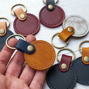 Op de afbeelding: Een collectie ronde lederen sleutelhangers in diverse kleuren, waaronder mosterdgeel, marineblauw, bordeauxrood en grijs. Elke sleutelhanger is voorzien van een bijpassende leren lus, een messingkleurige klinknagel en een ronde sleutelring.