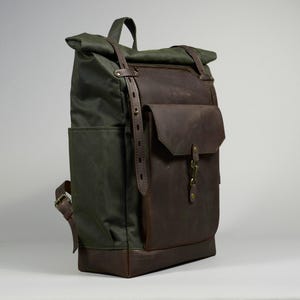 Könnte beinhalten: Ein Rolltop-Rucksack mit einem Korpus aus dunkelolivgrünem Canvas sowie einer Vordertasche, einem Boden und Besätzen aus dunkelbraunem Leder. Der Rucksack verfügt über verstellbare Lederriemen mit Metallschnallen und einen zentralen Metallverschluss.