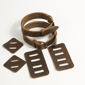 May include: Two brown leather straps with metal buckles and four brown leather patches. Two patches are square with two horizontal slots, and two patches are rectangular with four horizontal slots, all featuring visible stitching along the edges.